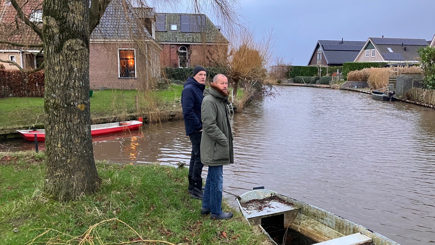 Two people in jackets stand by a small canal with a rowboat nearby. Houses and trees line the opposite bank. The scene appears calm and overcast, with grass and bare branches in the foreground.