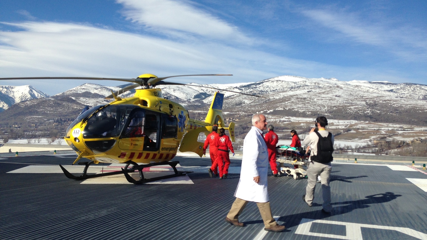 A yellow rescue helicopter on a helipad surrounded by snowy mountains. Several people, including a doctor in a white coat and others in red suits, are preparing to transfer a patient on a stretcher. The sky is clear and blue.