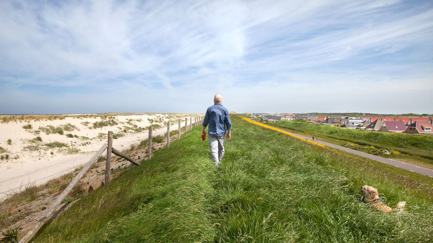 A person walks along a grassy path on a sunny day, with sand dunes on the left and a small town in the distance on the right. A dog lies in the grass nearby. The sky is partly cloudy.
