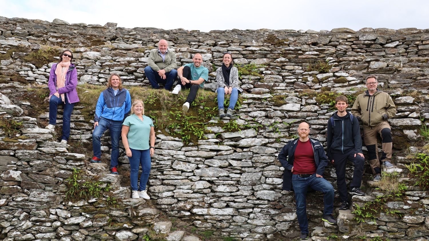 Nine people pose on the steps of an old stone ruin outdoors, sitting or standing at different levels. Some are smiling and dressed casually, with greenery growing between the rocks. The sky is partly cloudy.