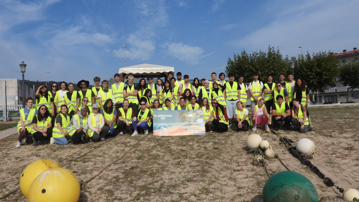 A large group of people wearing yellow reflective vests pose outdoors on sandy ground, with scattered buoys in front and a banner in the center. Trees, buildings, and a blue sky are visible in the background.