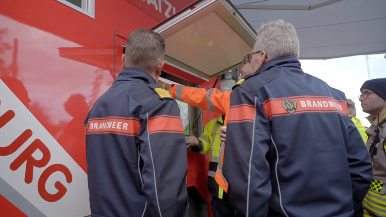 Several firefighters in navy uniforms with “Brandweer” on their backs stand near a red emergency vehicle, talking to personnel in yellow jackets. One person gestures toward a window on the vehicle.