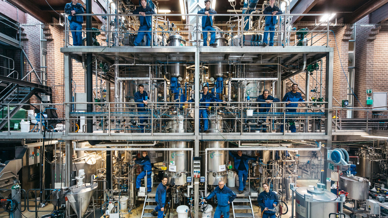 Several workers in blue uniforms stand on two levels of an industrial facility, surrounded by metal pipes, tanks, and machinery in a bright, spacious room with brick walls.