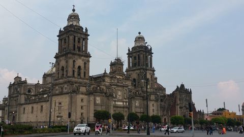 The image shows the Metropolitan Cathedral in Mexico City, a large historic stone building with two tall towers, intricate architectural details, and people walking in the plaza in front of it under a partly cloudy sky.