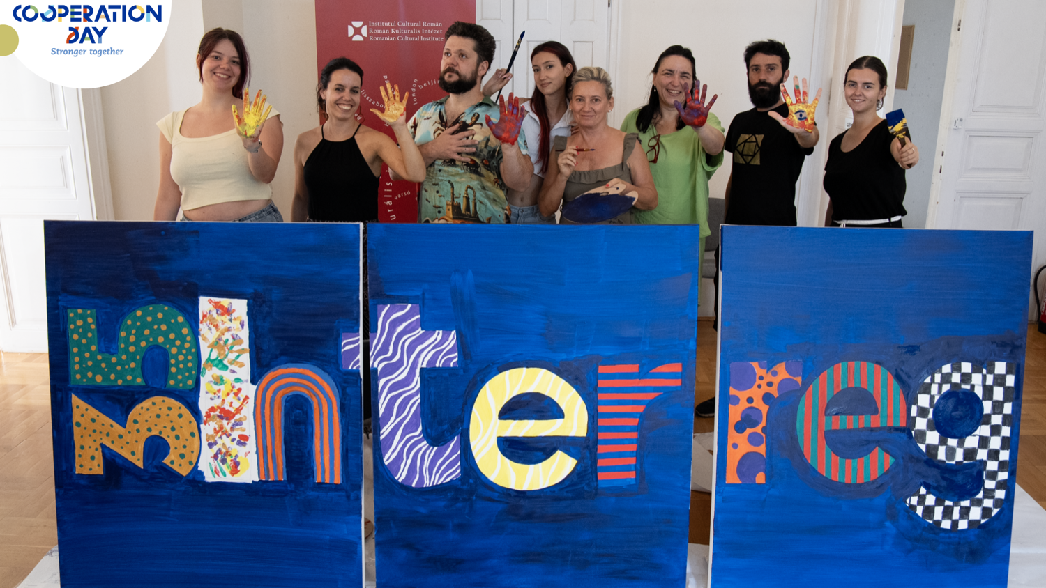 A group of nine smiling people stand behind three large painted panels spelling "Interreg," holding up their paint-covered hands. The Interreg Cooperation Day logo is in the top left corner.
