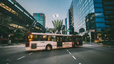 A red and white bus moves through a city intersection with modern glass buildings on either side. The street is empty and the sky is overcast, creating a calm urban atmosphere. Streetlights are on, adding a touch of illumination.