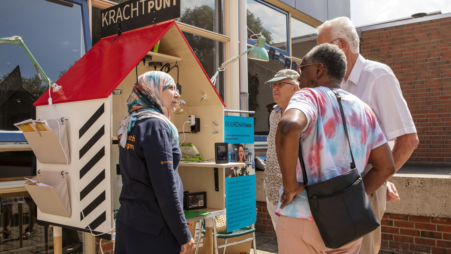 A woman in a headscarf speaks with three older adults near a small outdoor information booth called “KRACHTPUNT.” They are standing on a sunny sidewalk in front of a brick building.
