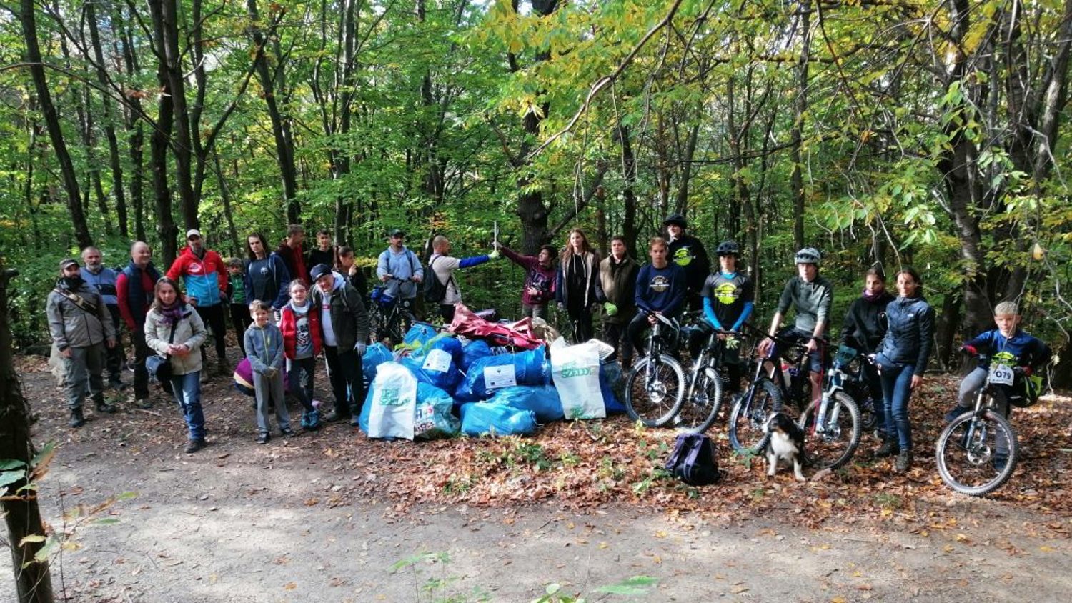 A group of people, including cyclists and a dog, stand in a forest clearing. They are gathered around numerous bags of collected litter. The scene suggests a community clean-up event in an autumn forest.