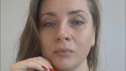A woman with long brown hair and blue eyes, wearing a green top, rests her chin on her hand with red-painted nails. She looks directly at the camera with a neutral expression against a plain background.