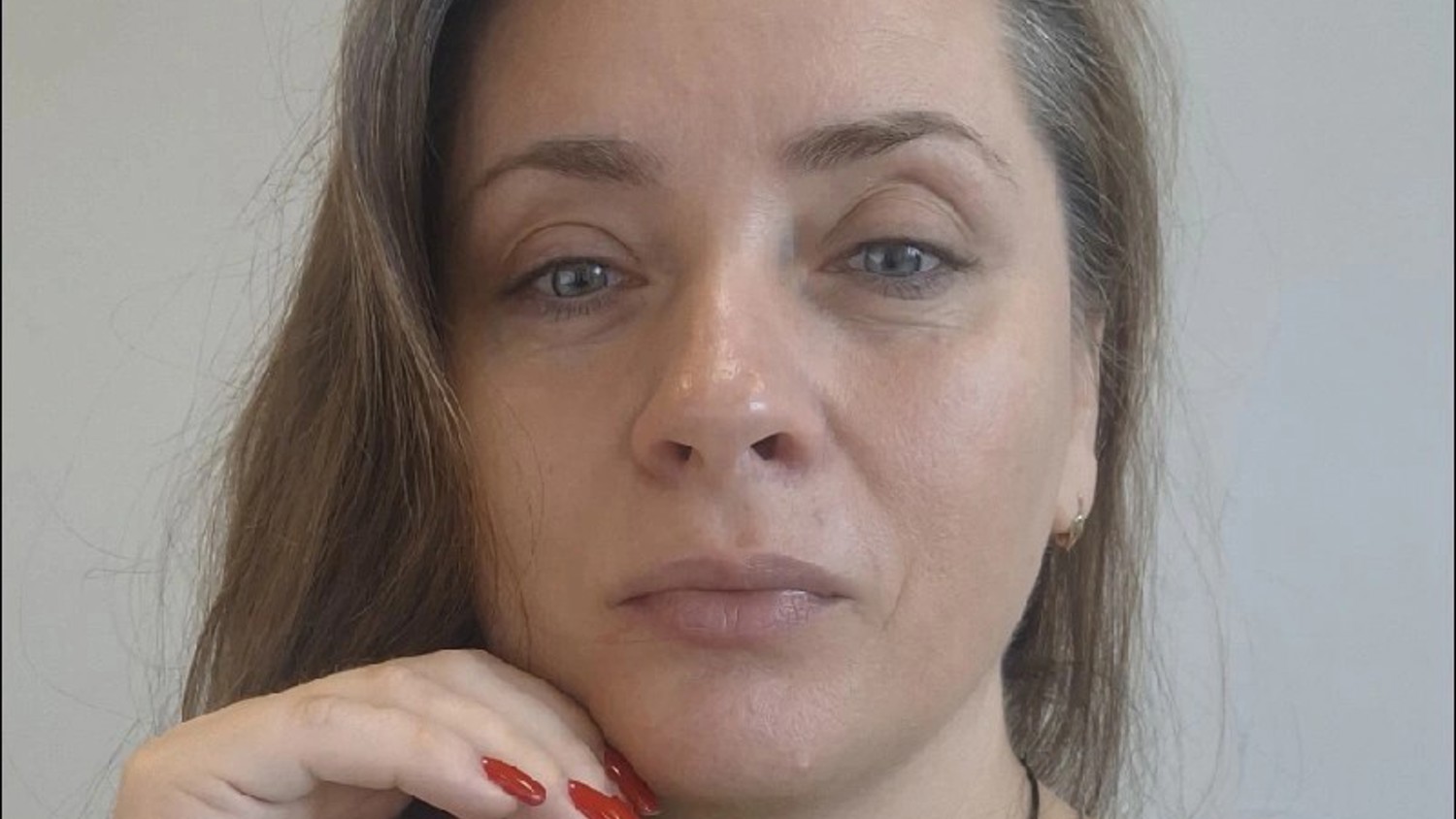 A woman with long brown hair and blue eyes, wearing a green top, rests her chin on her hand with red-painted nails. She looks directly at the camera with a neutral expression against a plain background.