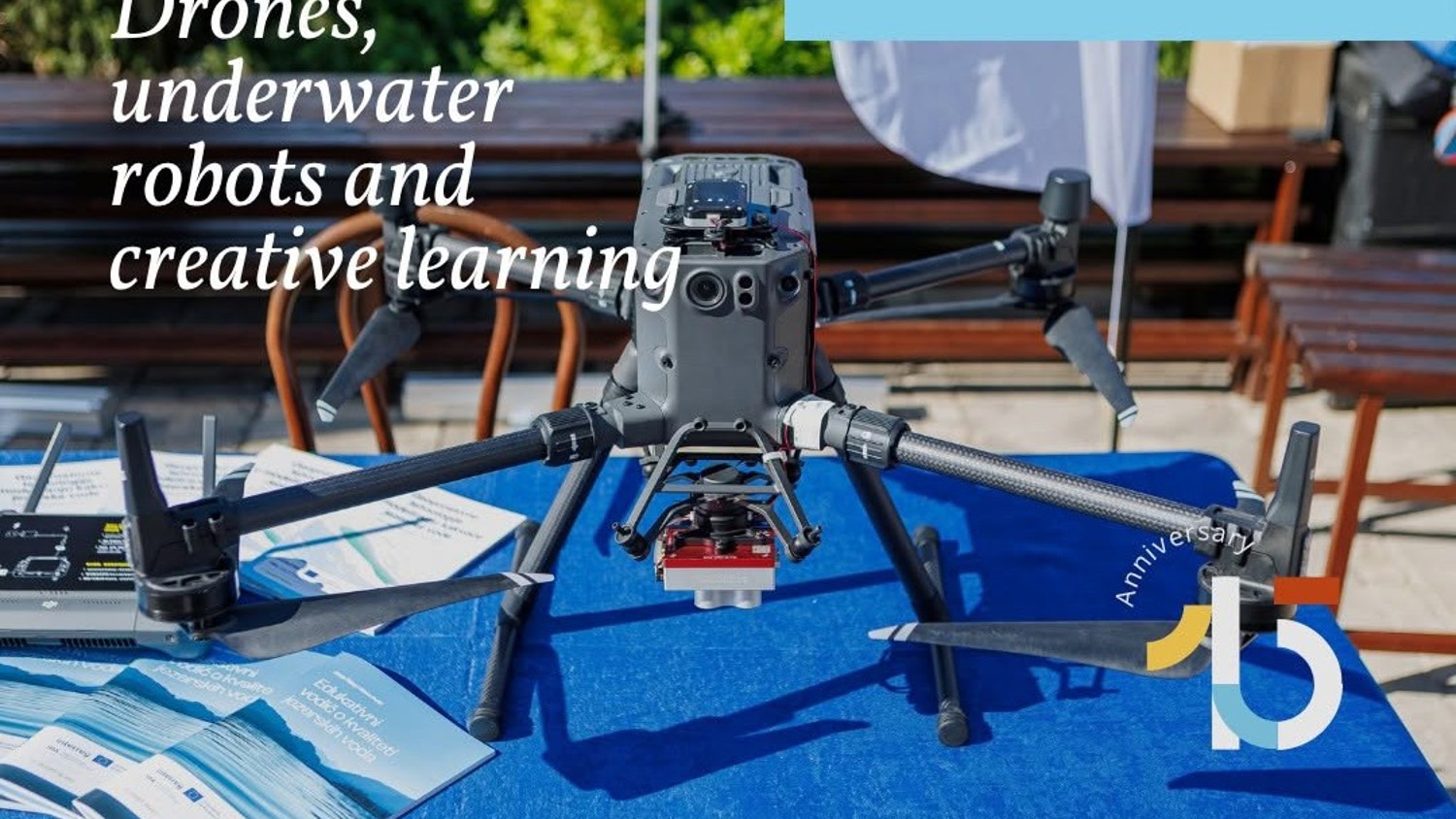 A drone is displayed on a blue table outdoors, surrounded by brochures. Text reads, “Drones, underwater robots and creative learning.” A partial anniversary logo appears on the lower right.