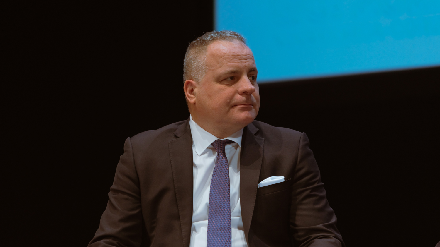 A man in a dark suit and blue tie sits holding papers, looking to his left. He is in front of a dark background with a blue screen above him.