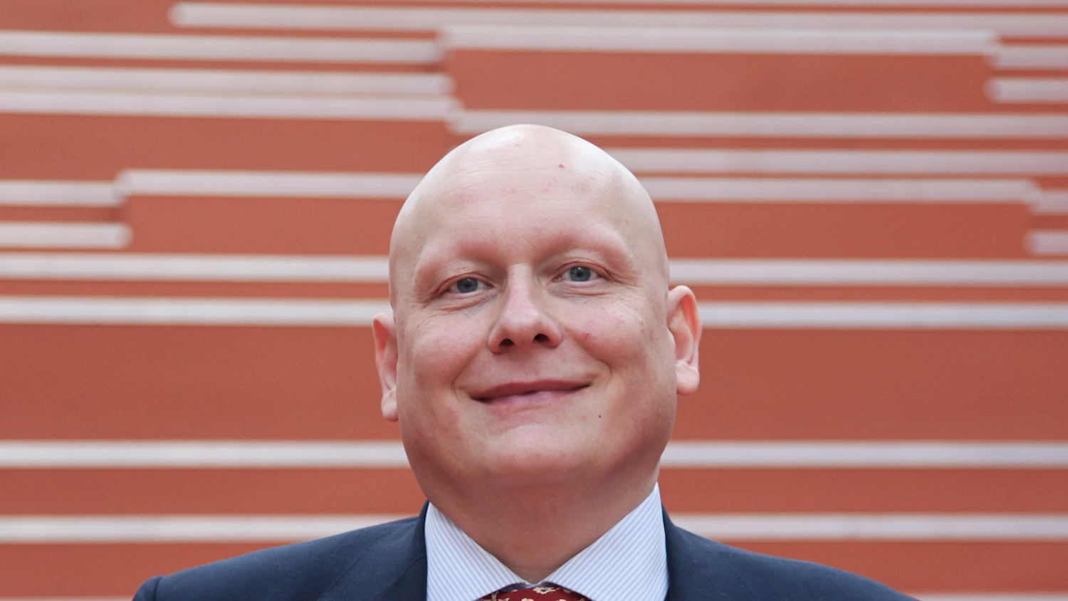 A person in a formal suit, with a pink pocket square and a patterned red tie, stands smiling in front of red stairs.