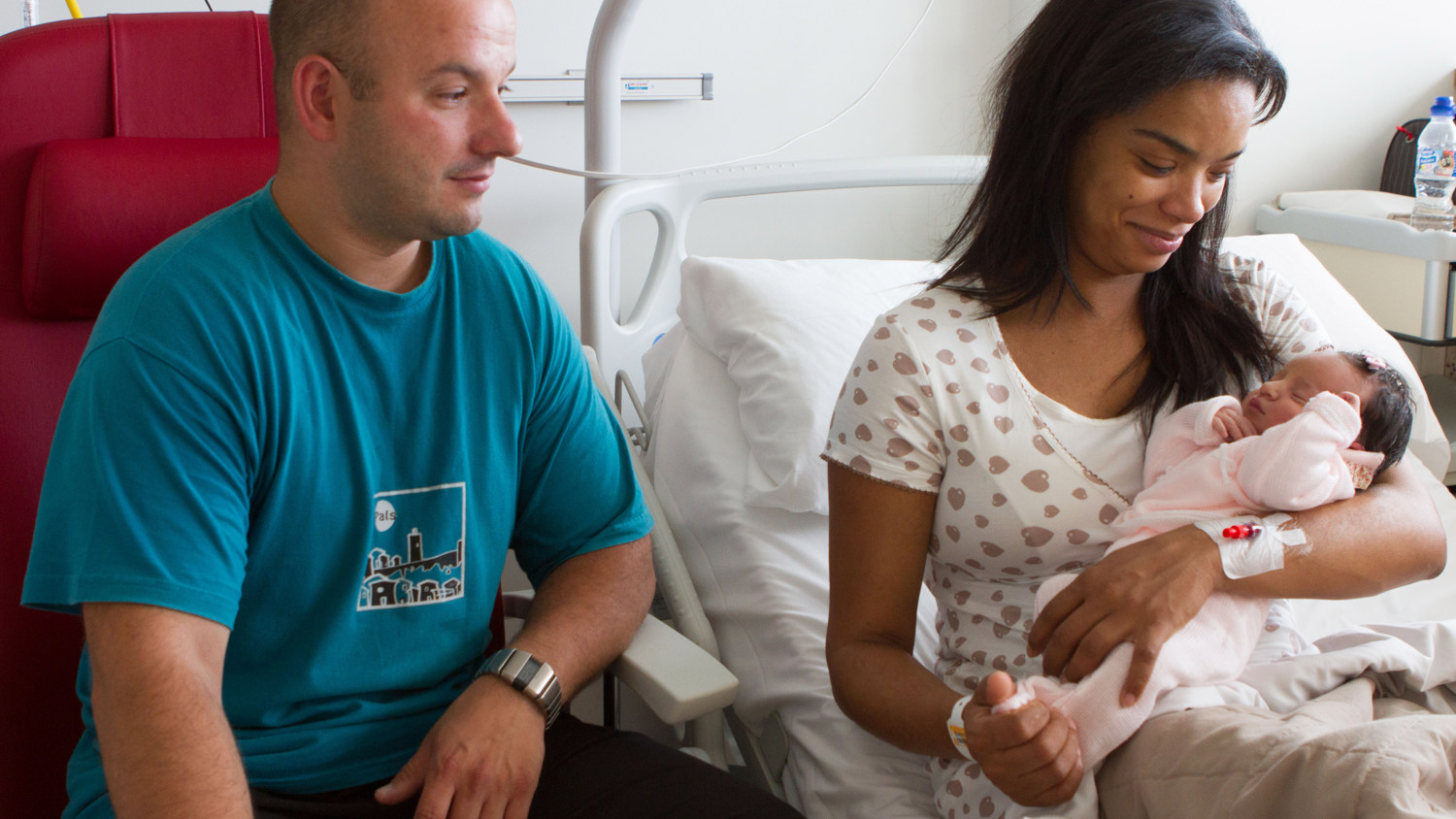 A woman sits on a hospital bed, cradling a newborn baby in her arms. She is wearing a hospital gown and has an IV attached to her hand. A man sits nearby in a red chair, wearing a blue t-shirt, and looks at the baby with a smile.