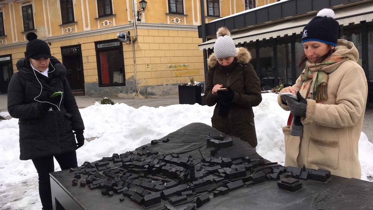 Three women in winter clothing look at their phones while standing around a tactile city model outdoors. Snow covers the ground, and historic buildings are visible in the background.