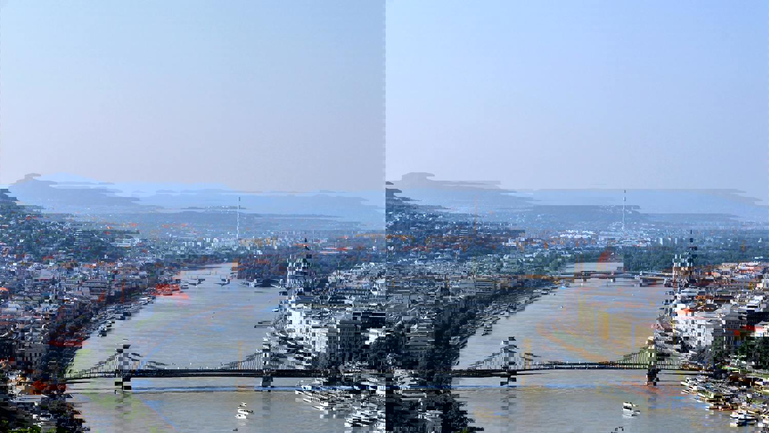 A cityscape featuring a wide river crossed by several bridges, with buildings and greenery on both banks. The image is distorted with digital glitches and pixelation, creating a broken, fragmented effect.