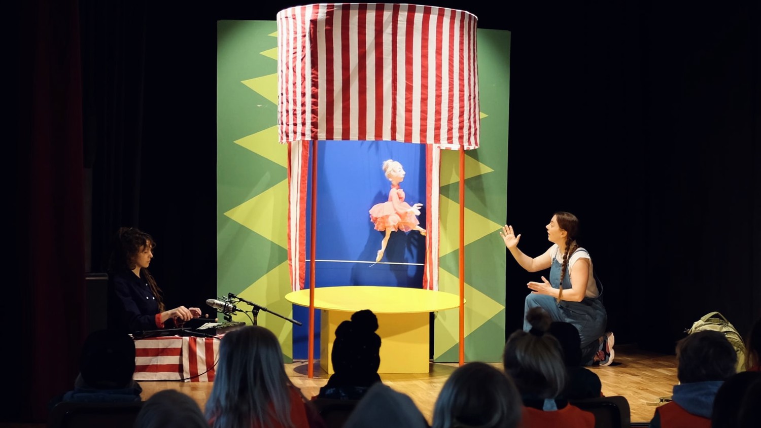 A woman performs as a puppeteer on stage with a red and white striped booth and a colorful backdrop. She kneels, interacting with a puppet in a pink dress. An audience watches from the foreground.