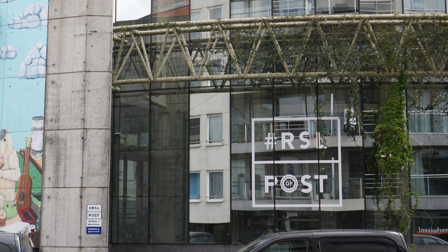 Glass-walled building with “#RS|POST” written on the window. A mural with people and clouds is painted on the left wall. Overhead, a vine-covered walkway connects buildings. Cars are parked in front.