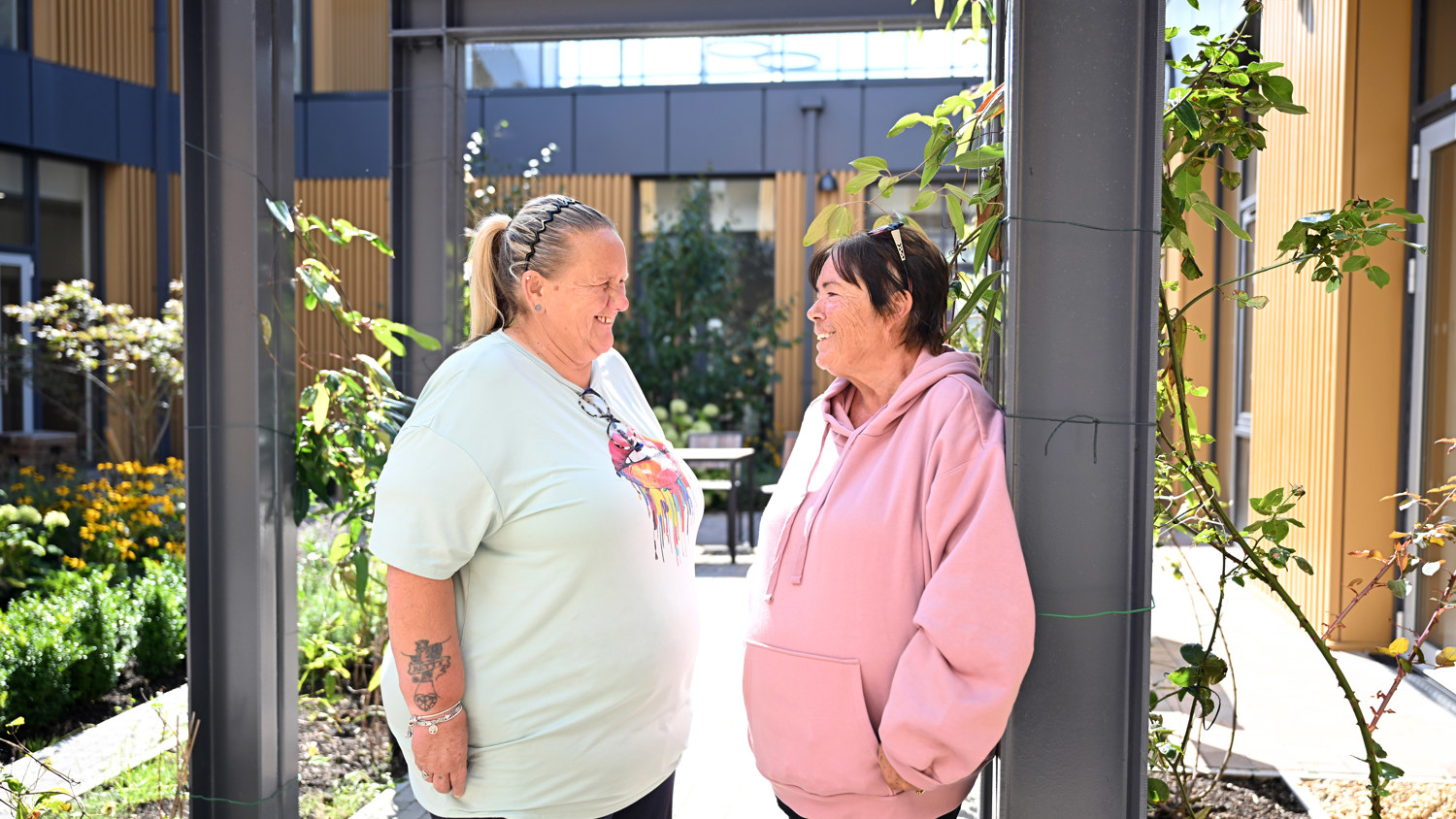 Two women stand outside in a garden, facing each other and smiling. One wears a light blue shirt and the other a pink hoodie. They are framed by metal beams and surrounded by plants, with a building in the background.