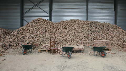 A large pile of bricks stacked in a warehouse with metal walls. In front, three wheelbarrows, a wooden chair, and two pallets with bricks are arranged on a dusty floor.
