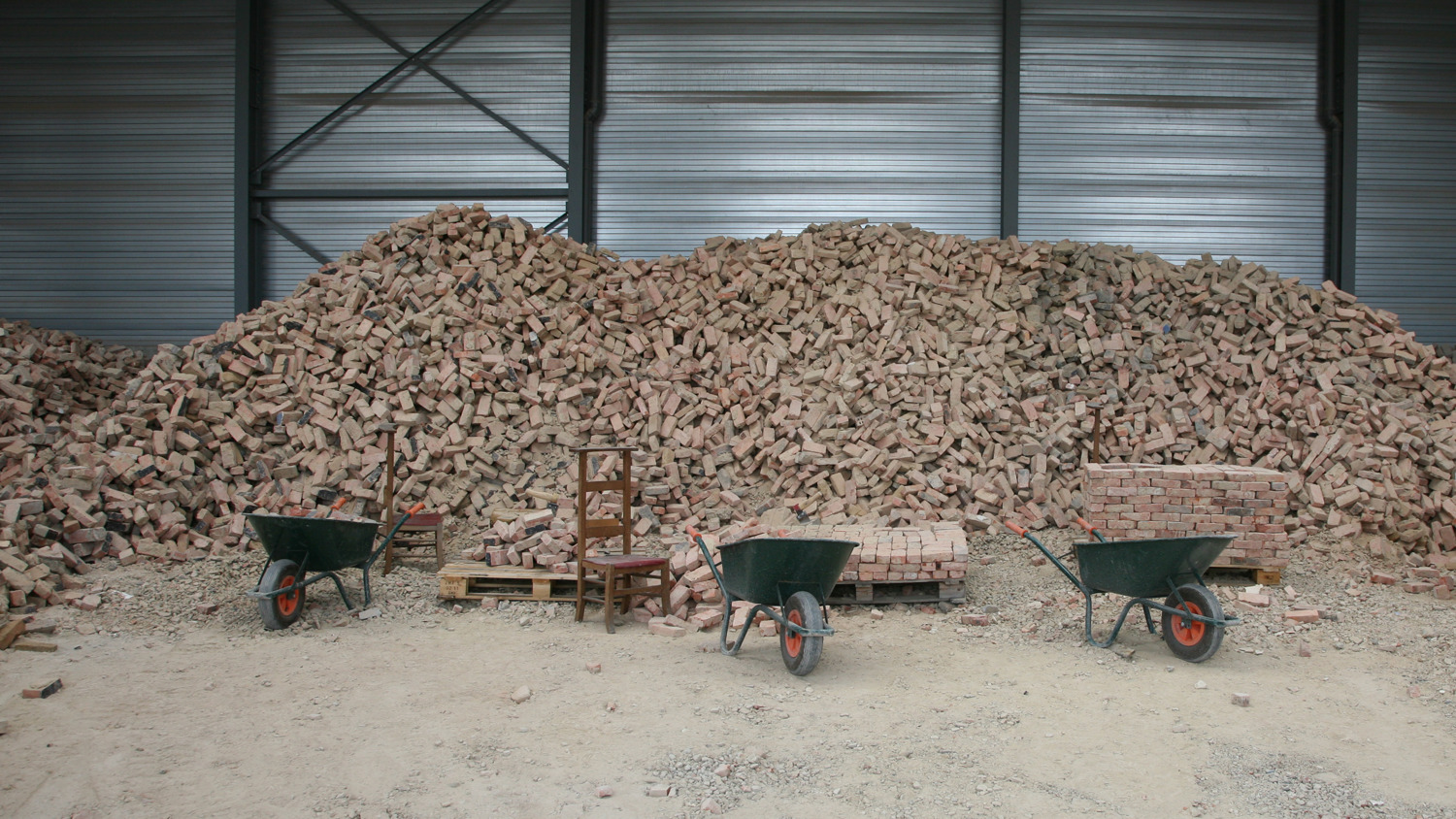 A large pile of bricks stacked in a warehouse with metal walls. In front, three wheelbarrows, a wooden chair, and two pallets with bricks are arranged on a dusty floor.