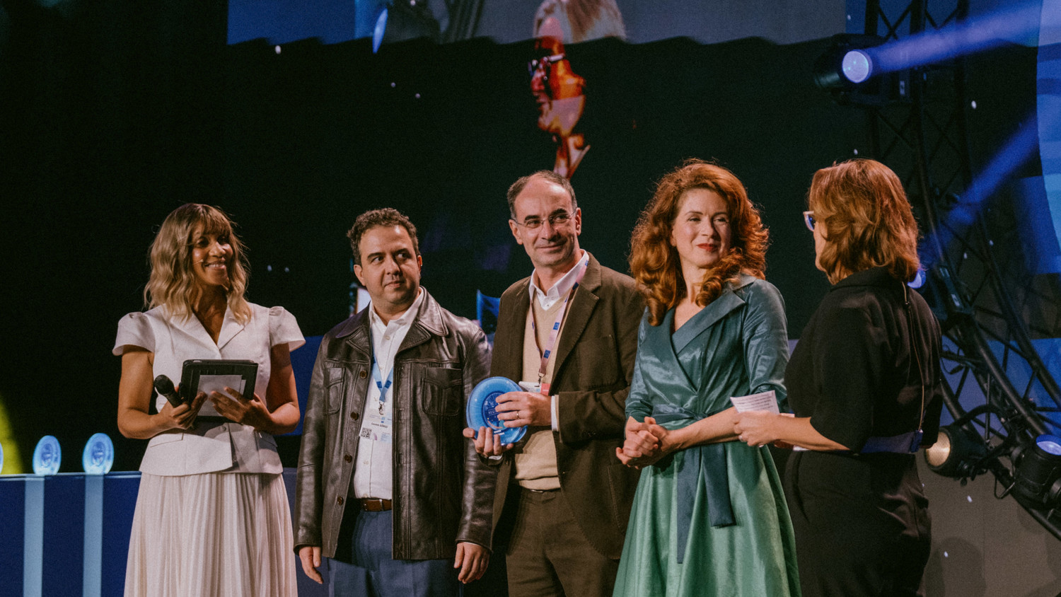 Five people stand on a stage under bright lights, some holding objects and papers. One woman wears a white dress, another a green dress, while the others wear business attire. An award ceremony appears to be taking place.