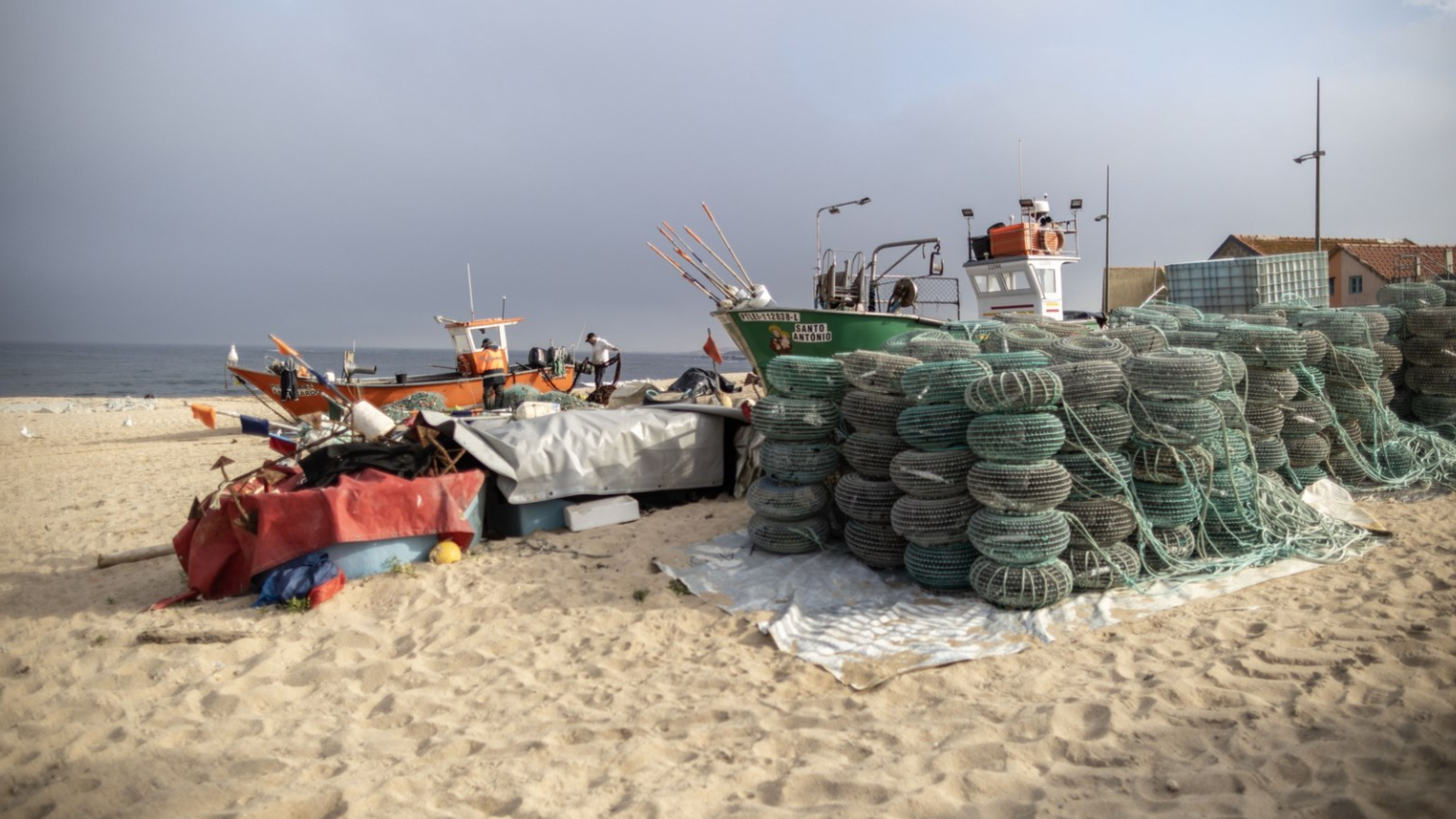 Fishing boats and large stacks of coiled nets or pots sit on a sandy beach near the ocean, with a few small buildings and a cloudy sky in the background.