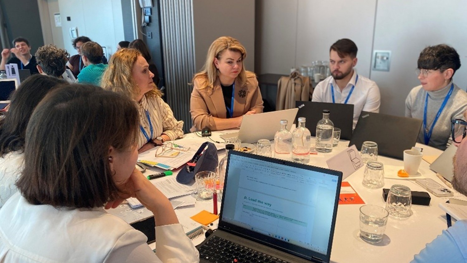 A group of people sit around a table in a conference room, engaged in discussion. Laptops, water bottles, and papers are spread out on the table. Some participants wear blue lanyards.