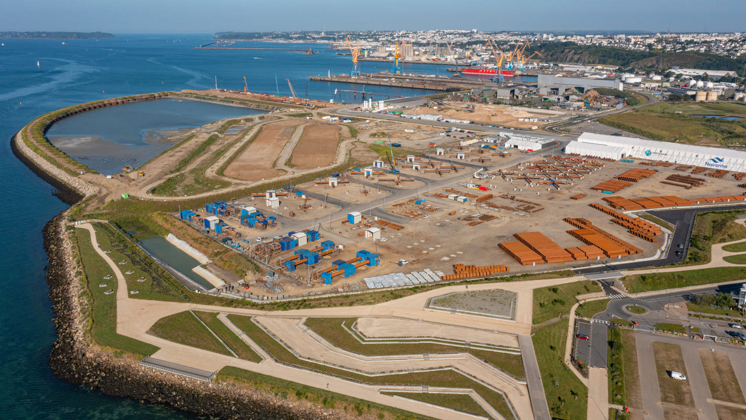 Aerial view of a large coastal industrial area with construction materials, machinery, and warehouses, surrounded by water and a breakwater, with a city and harbor visible in the background under a clear sky.