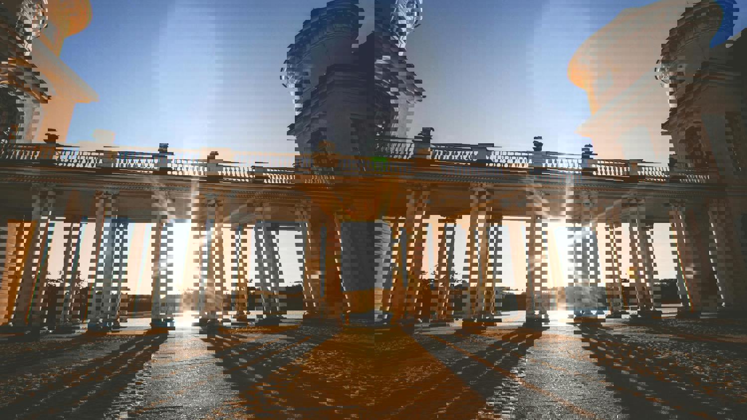 A pixelated, glitchy image of a colonnade with tall columns and sunlight streaming through, casting long shadows on the ground. The digital distortion affects the entire scene.