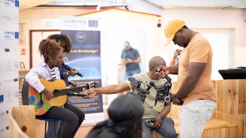 A young girl plays guitar and sings into a microphone while two cameramen film her. Another man in a yellow hat stands nearby, adjusting a camera. The setting appears to be an indoor event or interview.