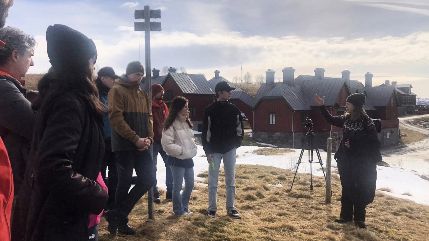 A group of people stands outside on patchy grass and snow, listening to a woman in a sweater who gestures while speaking. Red buildings with gray roofs are in the background under a cloudy sky.