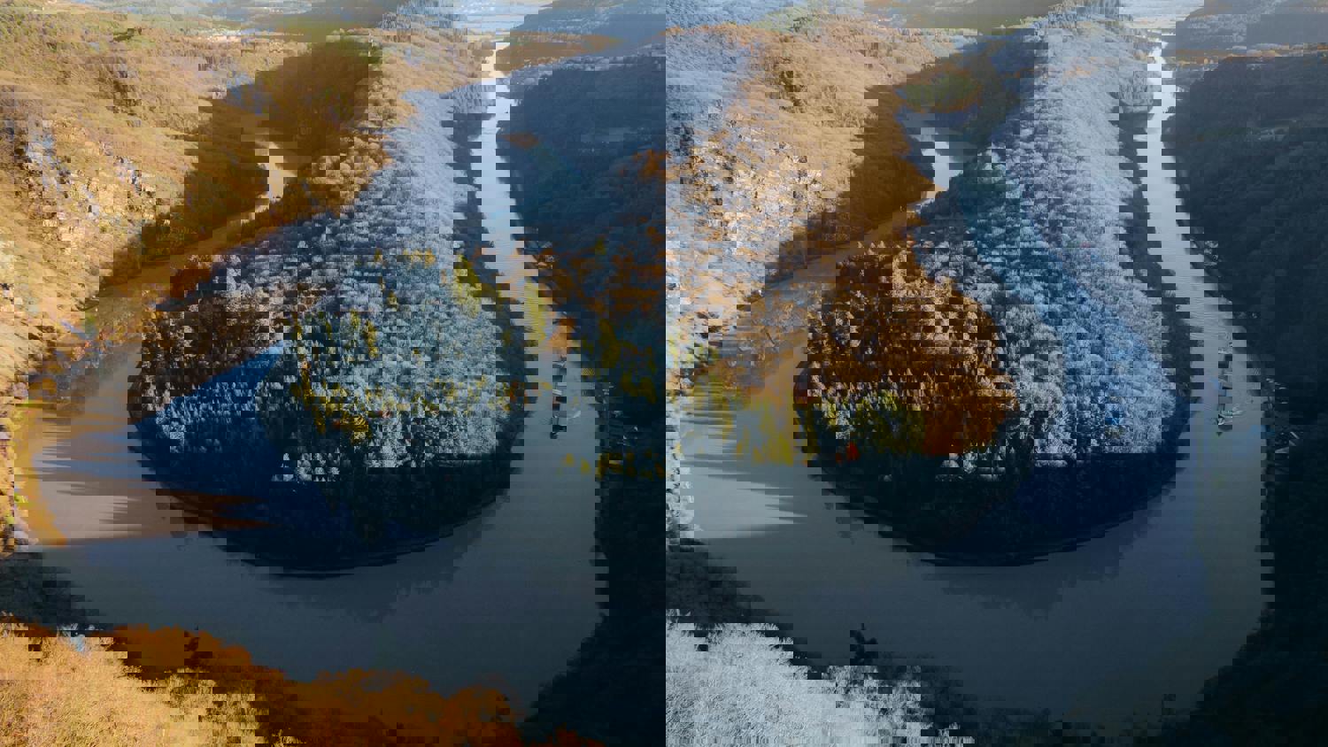 Aerial view of a winding river through forested hills, but the image is distorted with horizontal and vertical digital glitches, creating a pixelated and fragmented appearance.