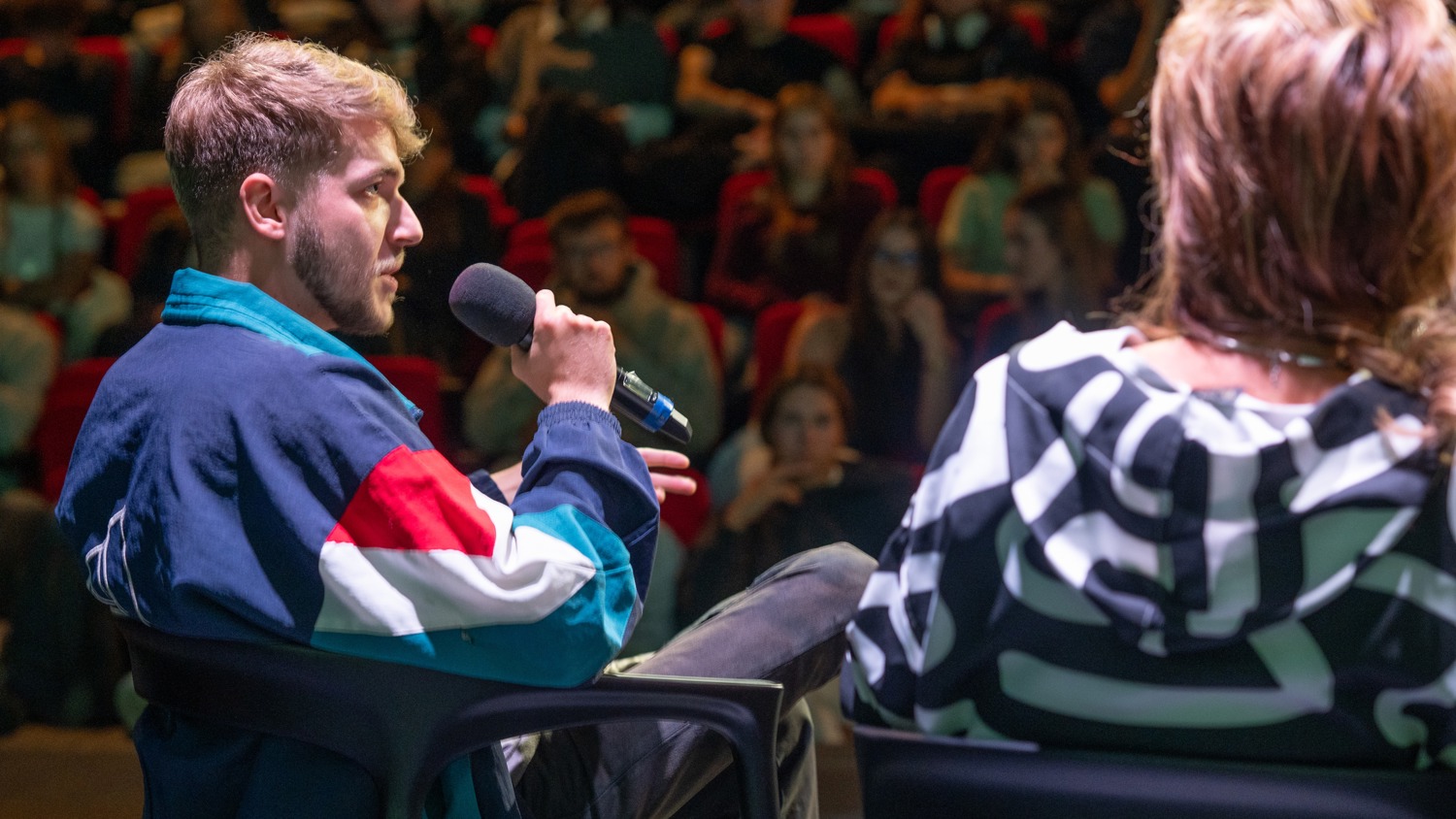 A man holding a microphone speaks while seated on stage next to another person, facing an audience in a large, dimly lit auditorium.