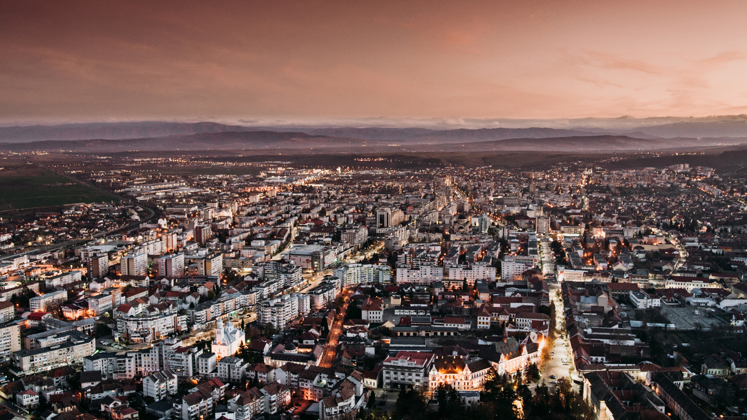 Aerial view of a city at dusk, with illuminated streets, densely packed buildings, and distant mountains under a dramatic, cloudy sky.