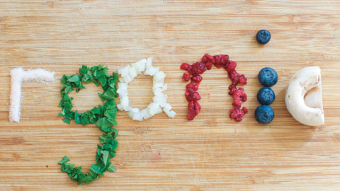 The word "organic" spelled out with food: an orange slice for "o," salt for "r," herbs for "g," chopped onion for "a," cranberries for "n," blueberries for "i," a mushroom slice for "c," and a wooden spoon with salt.