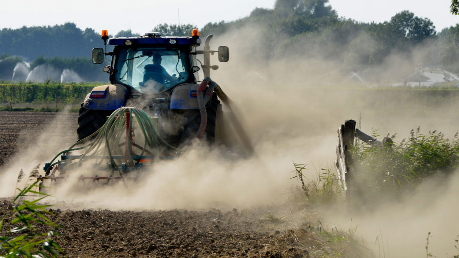 A blue tractor is plowing dry, dusty farmland surrounded by trees. Dust clouds rise behind the tractor as it moves forward, with fields and irrigation sprinklers visible in the background. The scene depicts rural agricultural work.