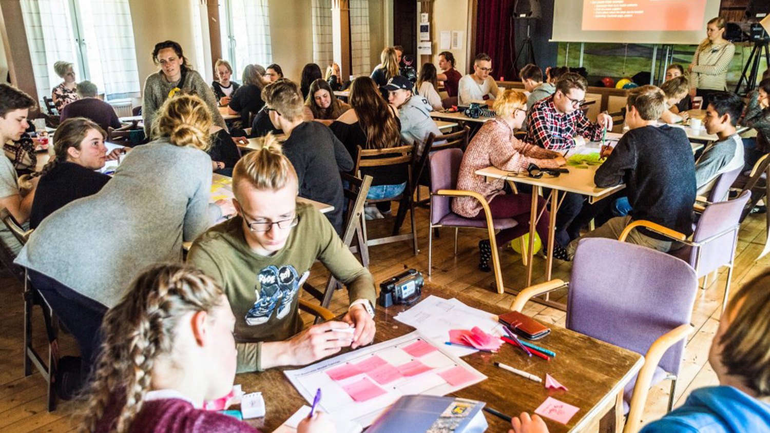A group of people seated around tables in a bright room, engaged in a collaborative workshop. There's a presentation on a red screen at the front of the room. Participants are discussing and writing on paper and sticky notes.