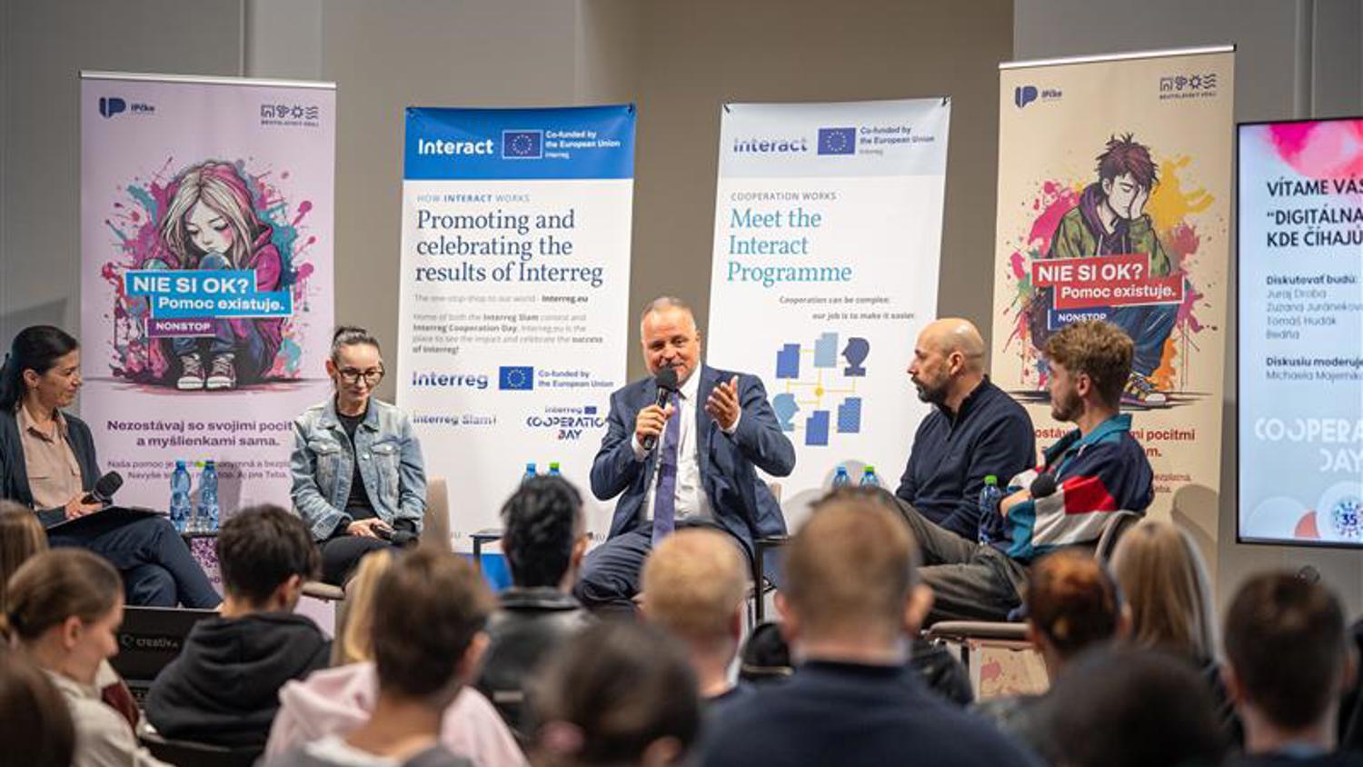 A panel of five people, including one man speaking into a microphone, sit on stage in front of an audience. Colorful banners and posters with text and graphics are displayed behind them.