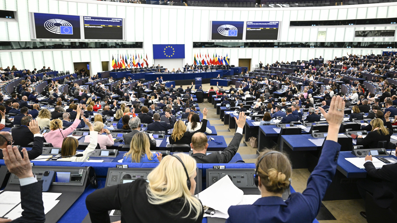 A large assembly of people seated in a modern parliamentary chamber, many raising their hands to vote. The European Union flag and member country flags are displayed at the front of the room.