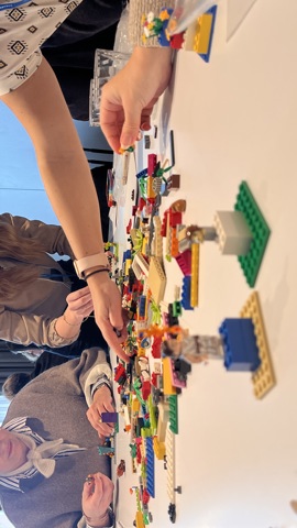 Four people sit around a table covered in colorful LEGO bricks, focused on building creations. One person is reaching for pieces, while others are assembling and examining their projects in a bright, modern room.