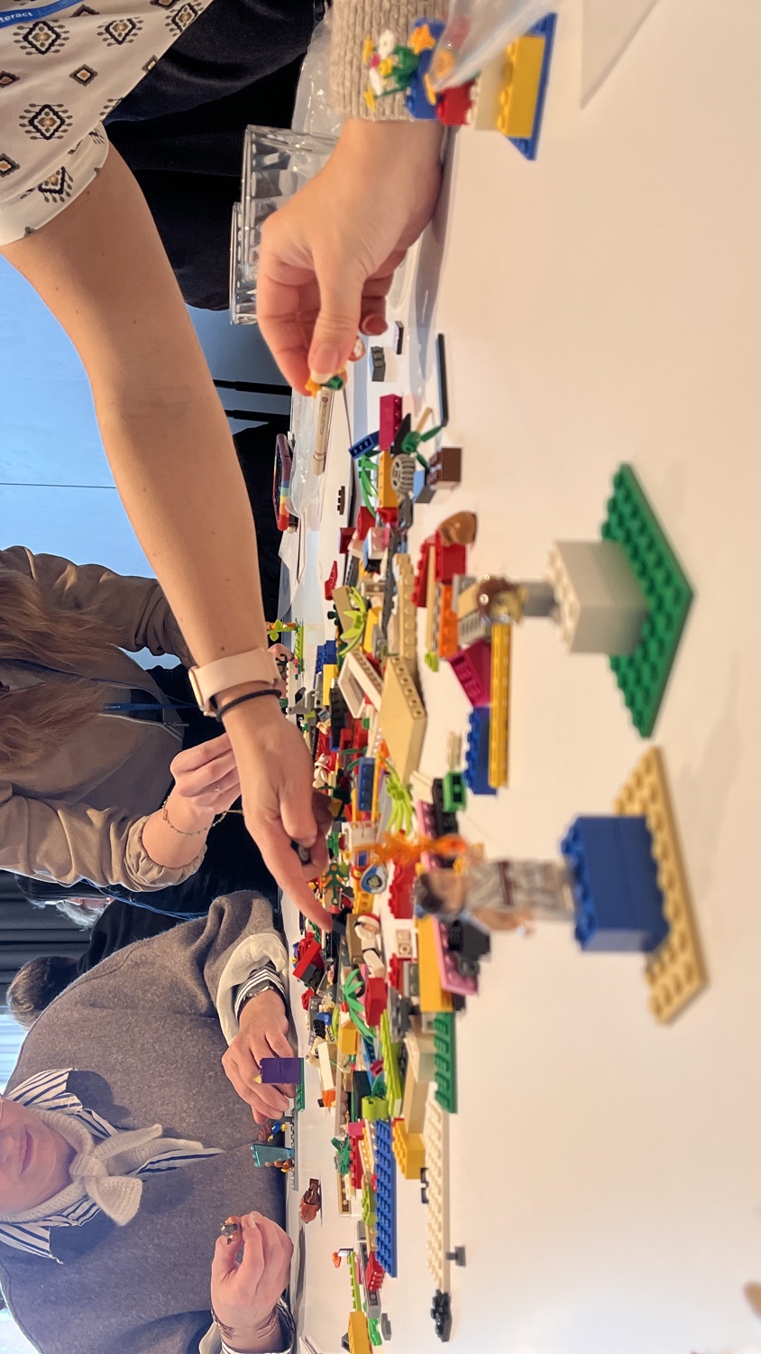 Four people sit around a table covered in colorful LEGO bricks, focused on building creations. One person is reaching for pieces, while others are assembling and examining their projects in a bright, modern room.