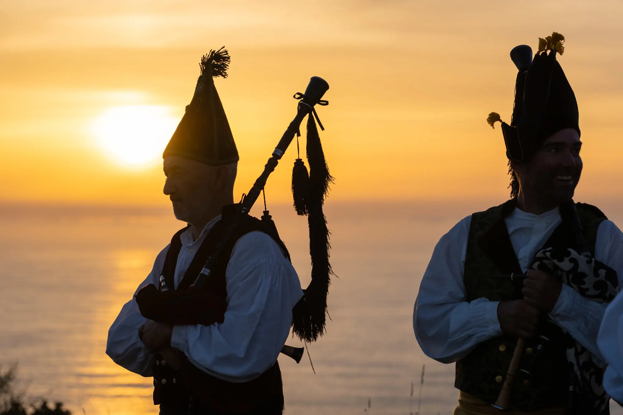 Two musicians in traditional clothing and tall hats play bagpipes at sunset, with the sun low over a calm sea in the background, casting a warm golden light.