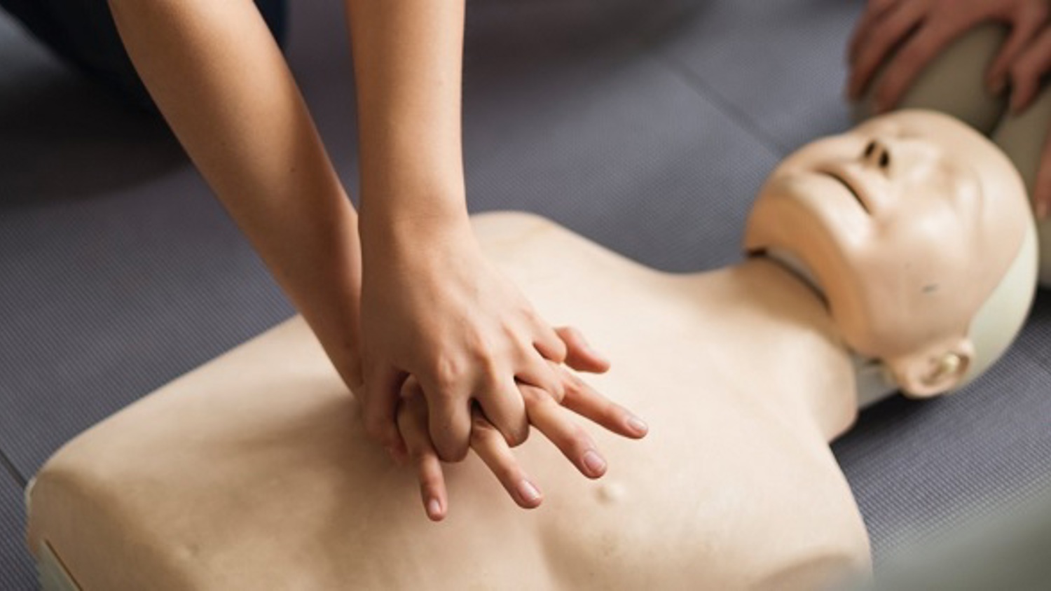 A person practices CPR on a dummy, with hands positioned on the chest. A CPR instruction sheet lies beside them on the mat.