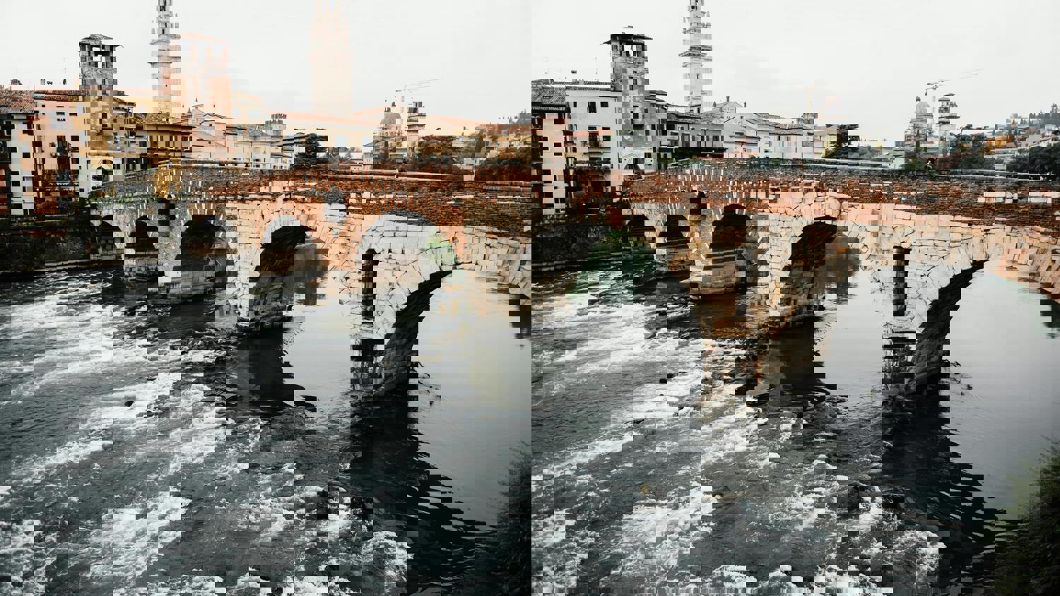 A distorted, pixelated image of an old stone bridge with multiple arches crossing a river, with buildings and cranes visible in the background. The water below appears wavy, and the scene is affected by glitch-like digital effects.