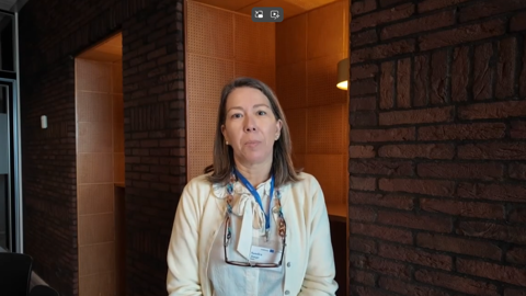 A woman with straight brown hair, wearing a cream cardigan, floral scarf, and name badge, stands indoors against a dark brick wall with a wooden alcove lit by a lamp behind her.