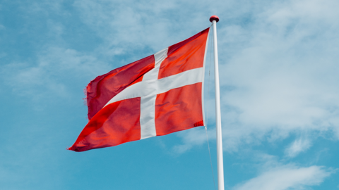 A red flag with a white Scandinavian cross, the national flag of Denmark, flies on a white flagpole against a blue sky with scattered clouds.
