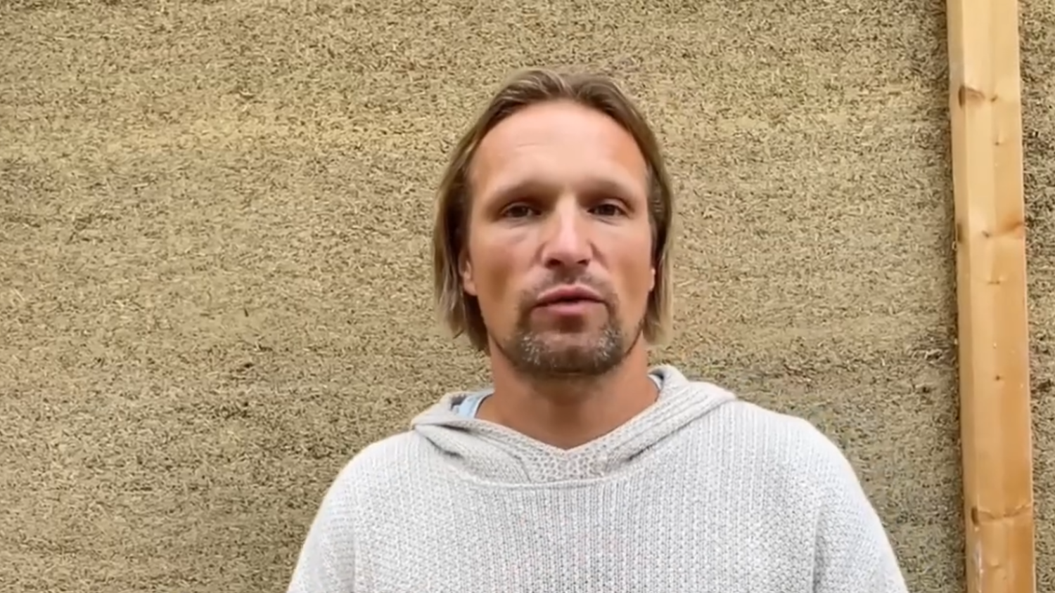 A person with light brown hair and a short beard, wearing a light gray knit sweater, stands in front of a textured beige wall with a wooden beam on the right.