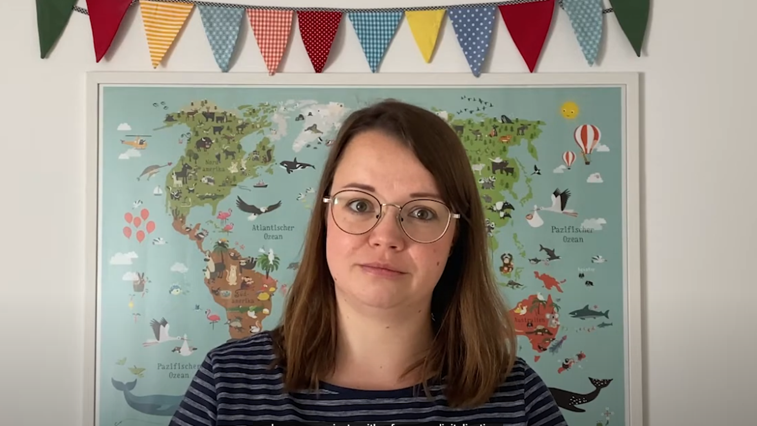 Woman with glasses and brown hair stands in front of a colorful world map. Above the map hangs a string of decorative, multicolored triangular flags. She is wearing a striped shirt and looking at the camera.