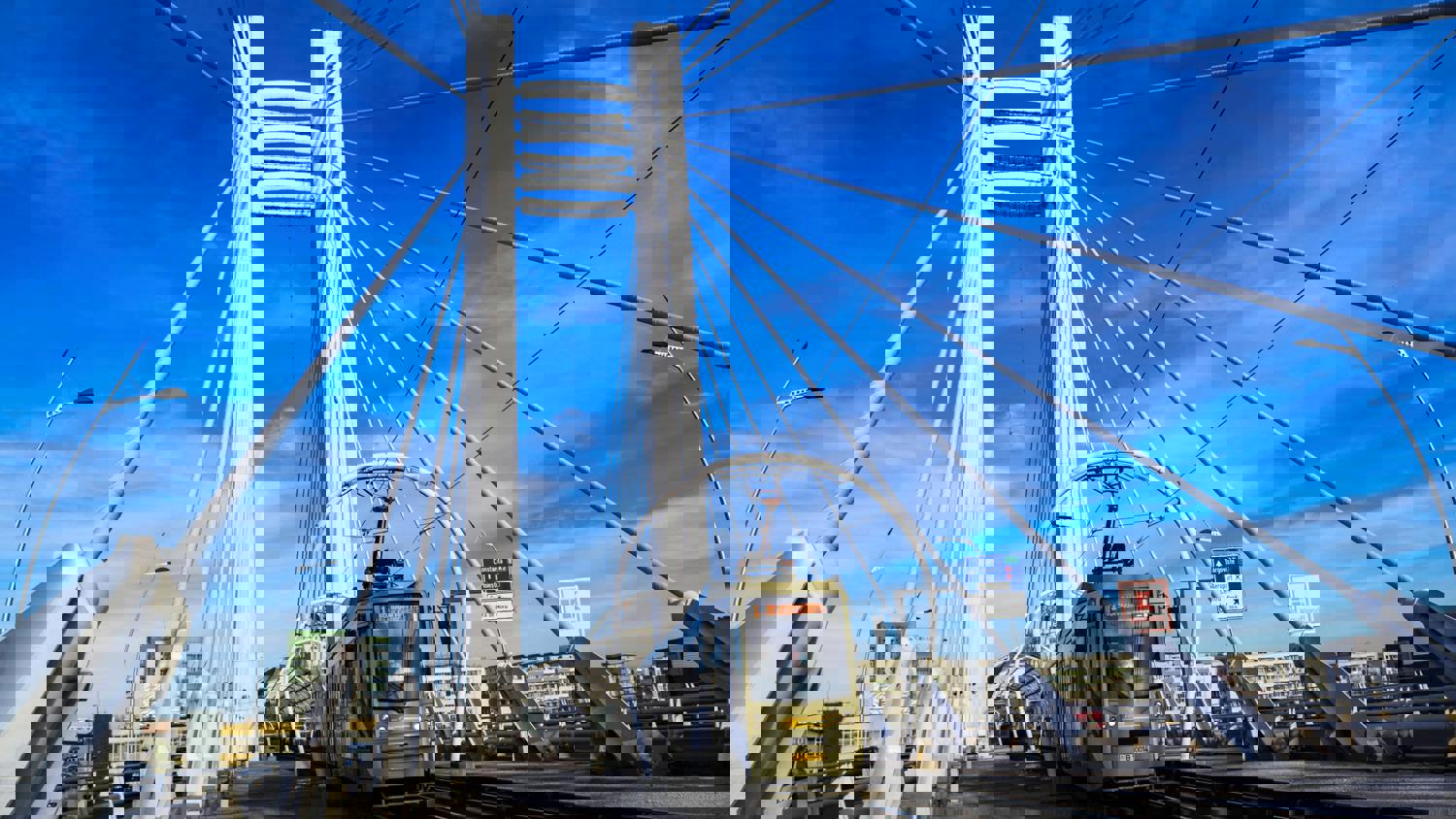 A city bridge with tall towers and suspension cables, set against a bright blue sky, appears heavily distorted with colorful digital glitches and pixelation.